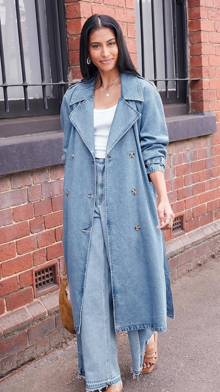 A woman posing in a long denim trench coat and light blue wide-leg jeans.