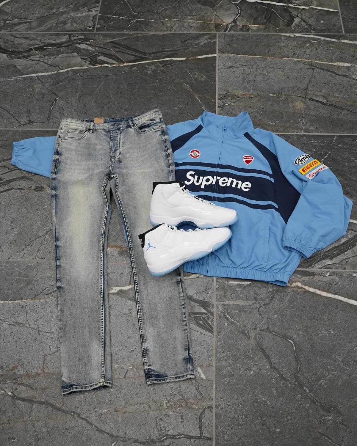 Light blue jeans, a Supreme track jacket, and white sneakers laid on grey tiles.