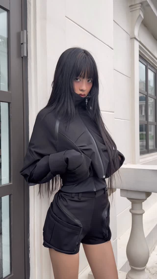 A woman with long black hair wearing a black cropped jacket and black shorts outdoors.