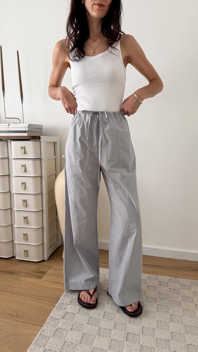 Woman wearing white tank top and high-waisted grey wide-leg trousers standing indoors.