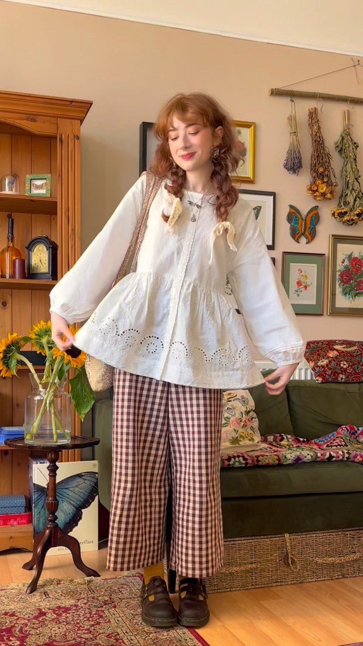 Woman wearing a long white eyelet blouse and checkered wide-leg trousers in a cozy room.