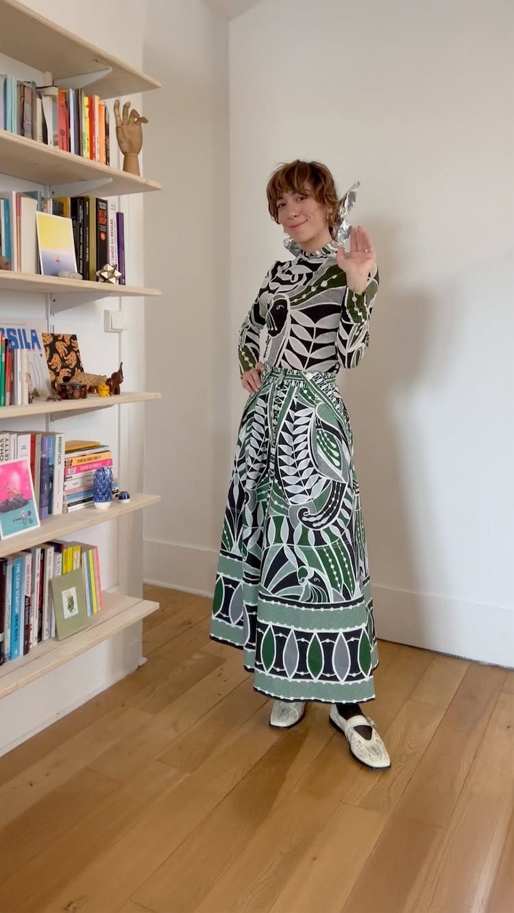 A woman wearing a long-sleeved green and black patterned maxi dress standing near bookshelves.