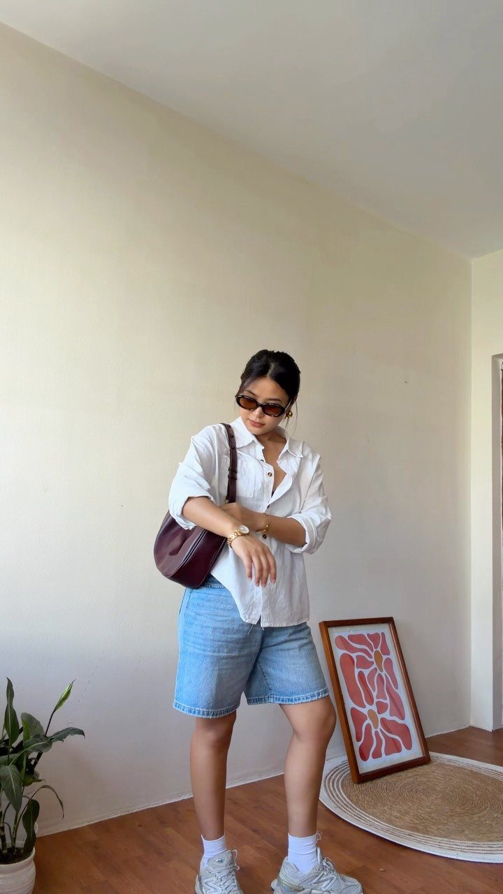 Woman posing in a white shirt and light blue denim shorts with white sneakers.