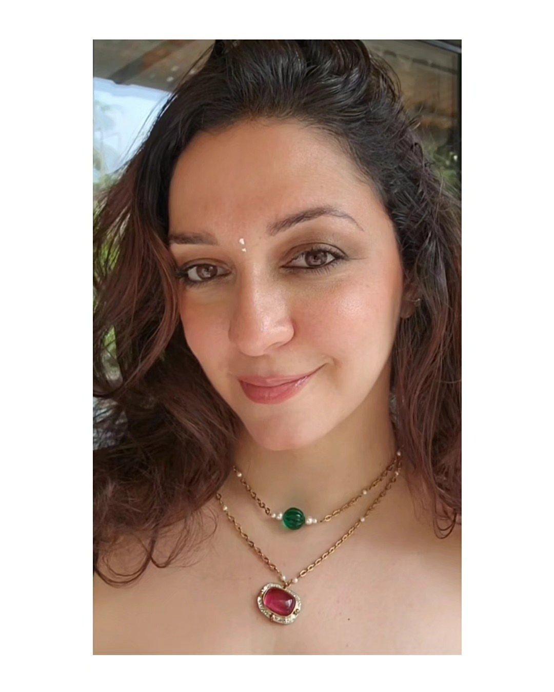 A woman wearing two gold layered necklaces with green and pink gemstone pendants.