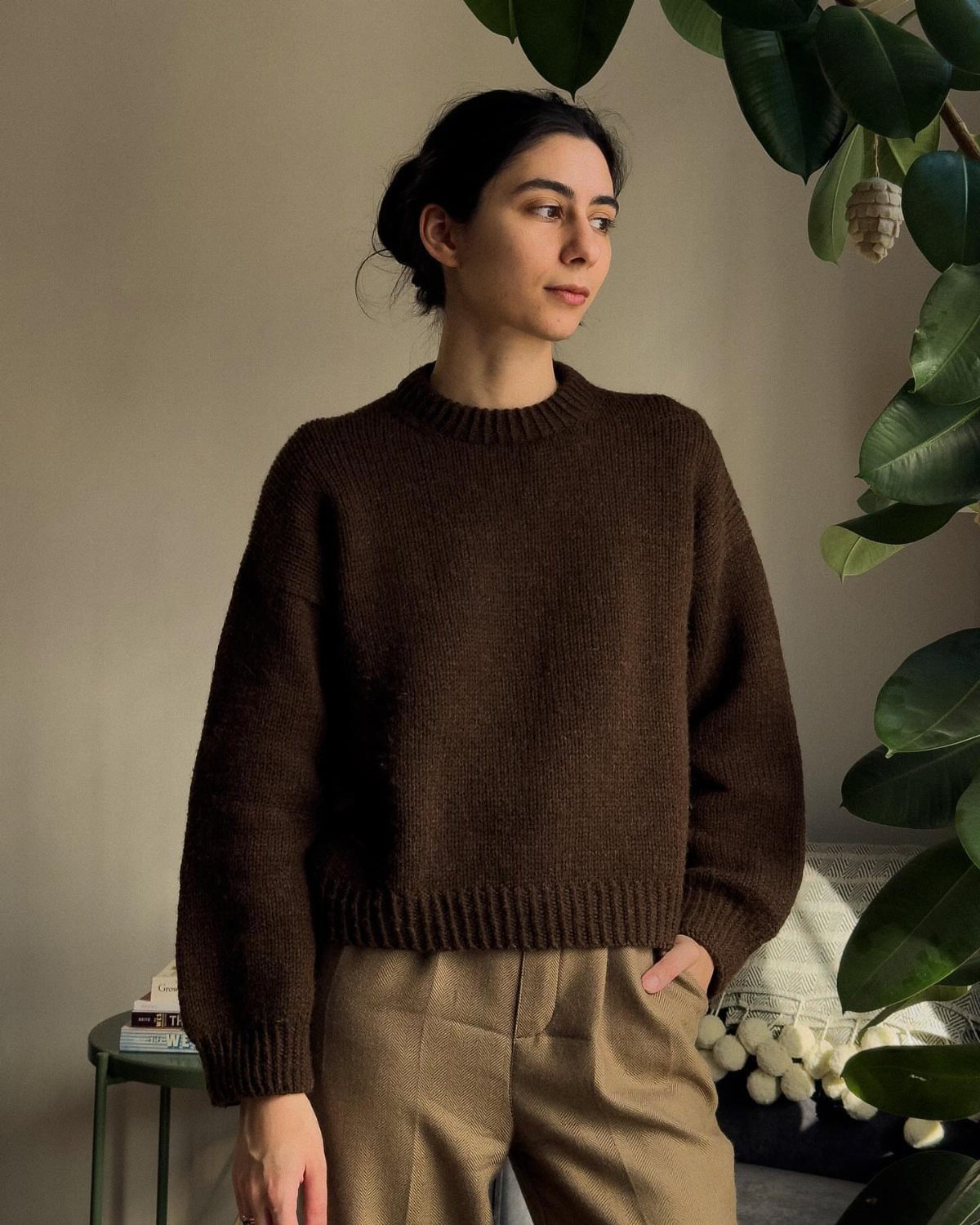 A woman wearing a dark brown chunky knit sweater and tan trousers standing near a plant.