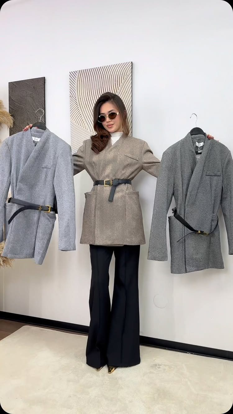 A woman posing in a beige blazer and black flared trousers between two hanging grey blazers.