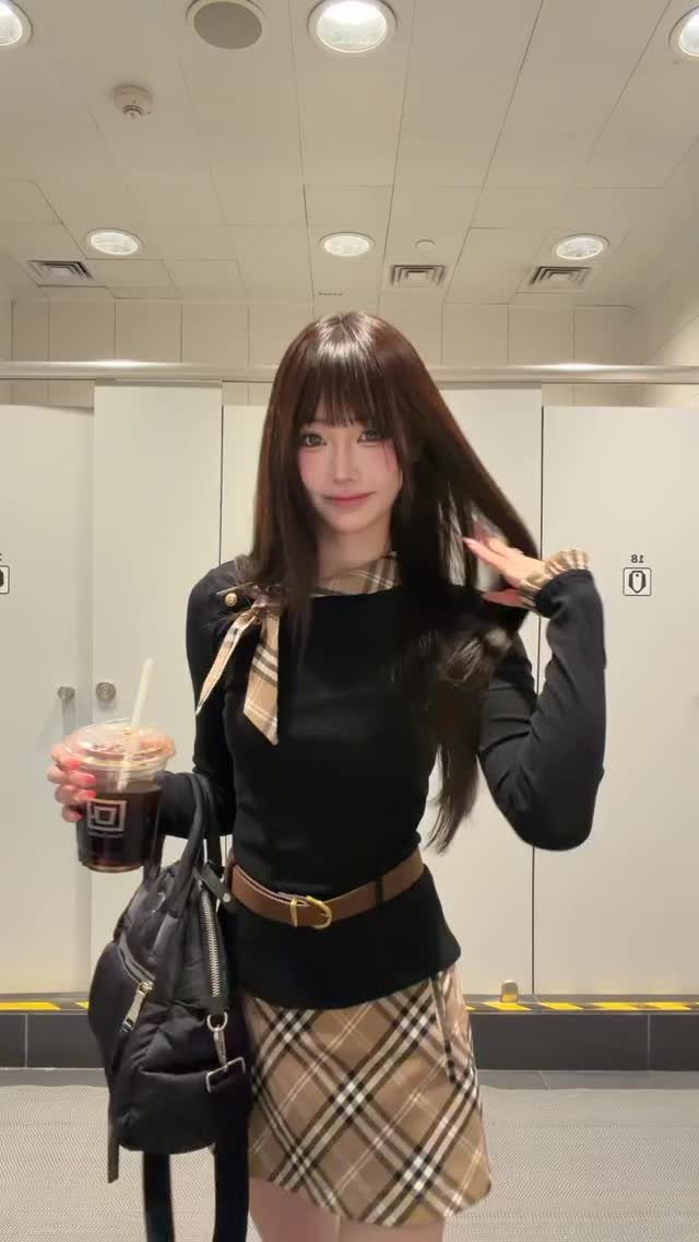 A young woman posing in a black top and a tan plaid mini skirt.