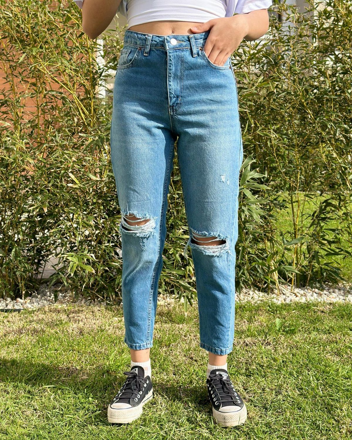 Person wearing distressed blue mom jeans, white crop top, and black canvas sneakers on grass.