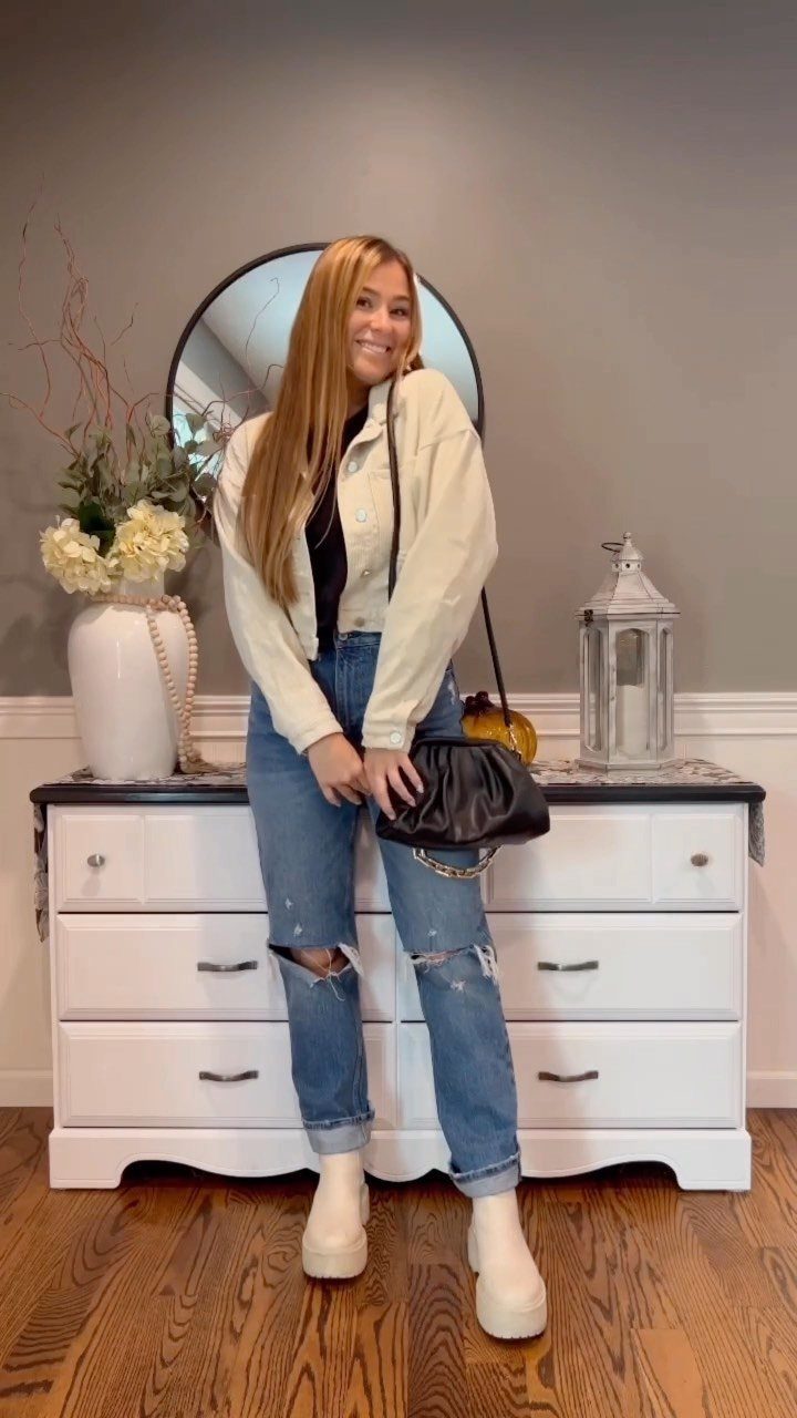 A woman posing in blue ripped mom jeans, a white cropped jacket, and white platform boots.