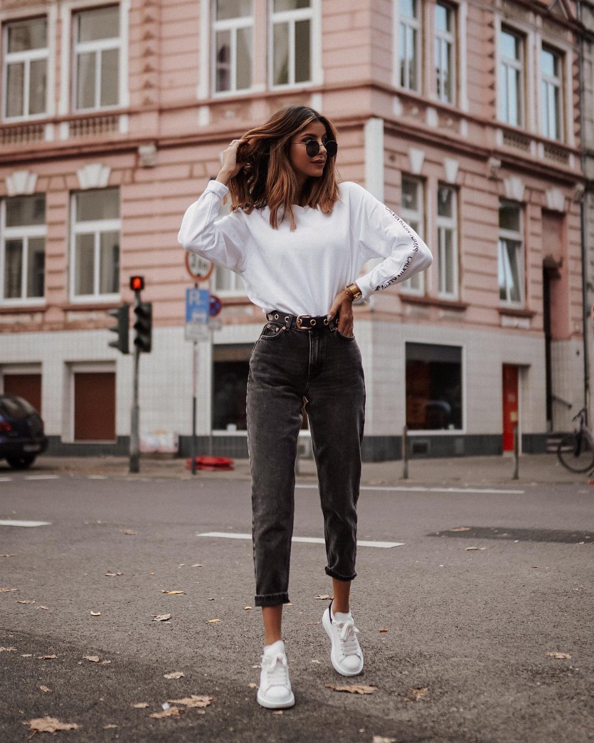 Woman in dark grey mom jeans, white sweatshirt, and white sneakers standing on a city street.