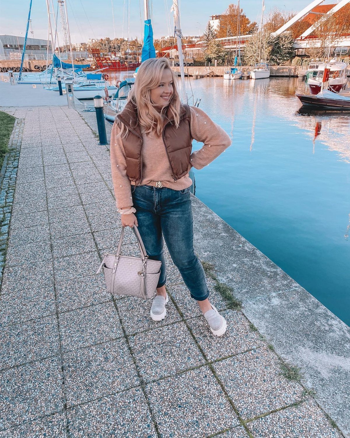 Blonde woman wearing blue mom jeans, a tan sweater, and a brown puffer vest by a harbor.