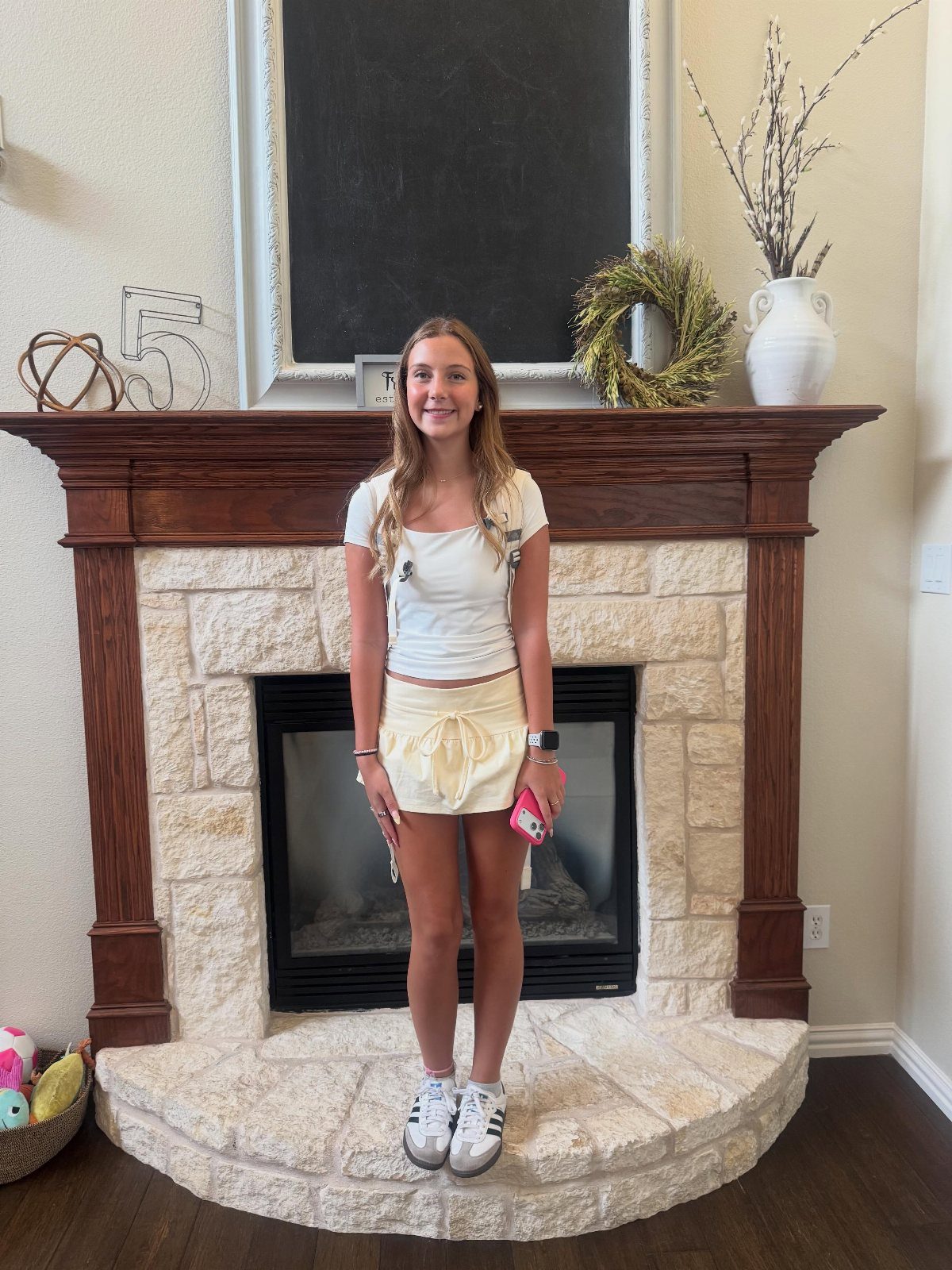 A smiling teenage girl wearing a white crop top, yellow mini skirt, and white sneakers standing by a fireplace.