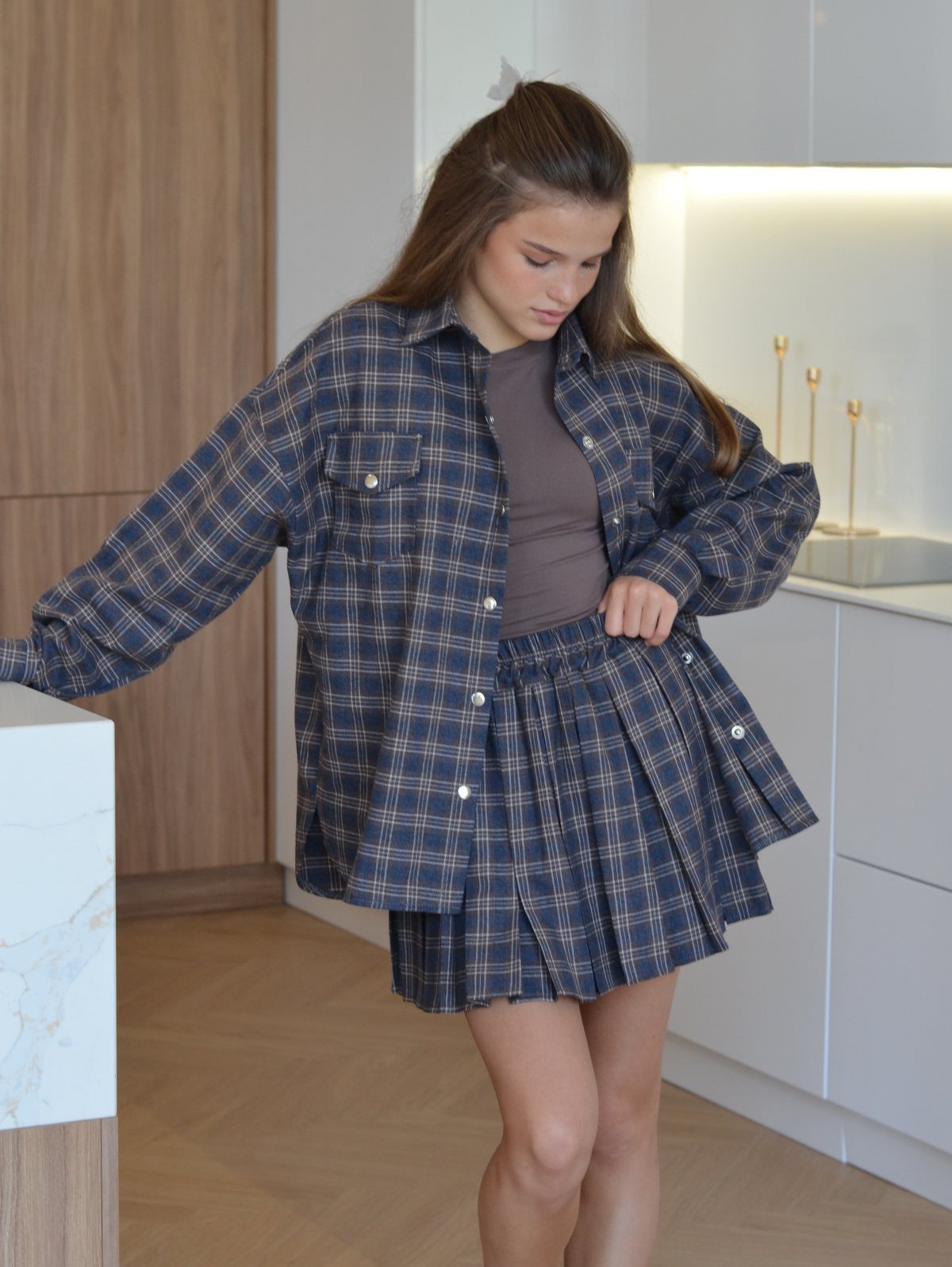 A young woman wearing a blue plaid oversized shirt and matching pleated skirt in a kitchen.