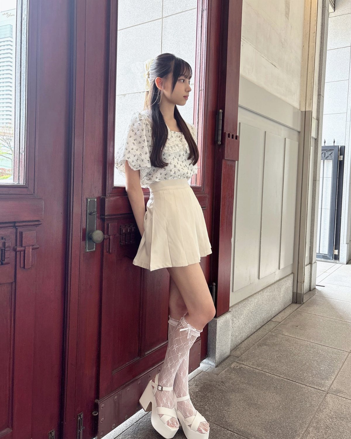 Young woman in a floral top, cream skirt, lace stockings, and white platform sandals leaning against a door.