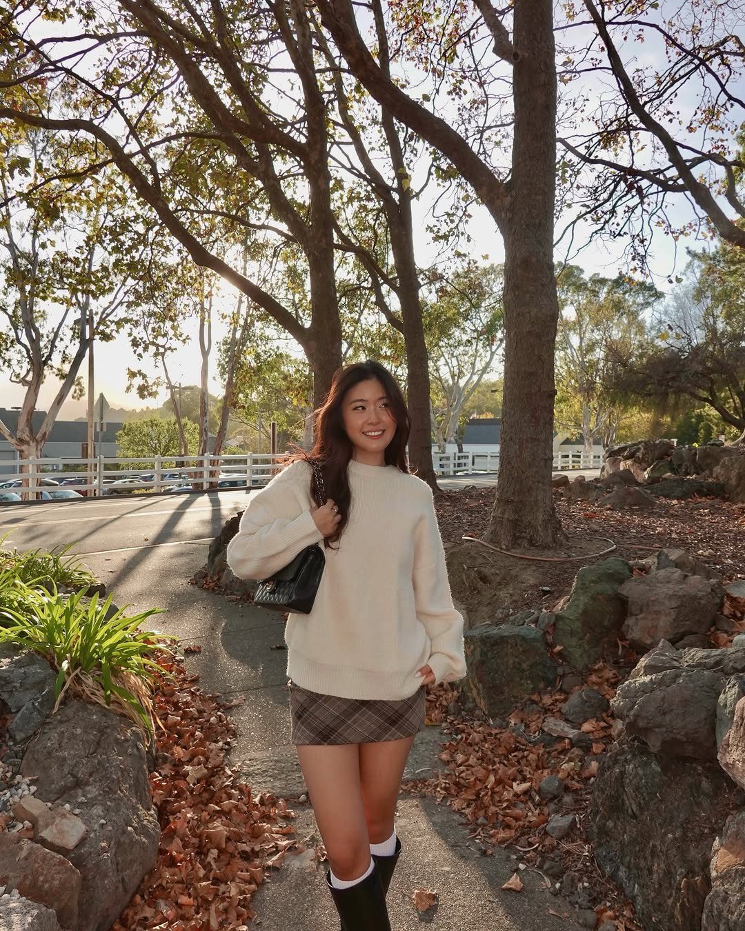 A smiling woman wearing an oversized cream sweater and plaid skirt standing outdoors.