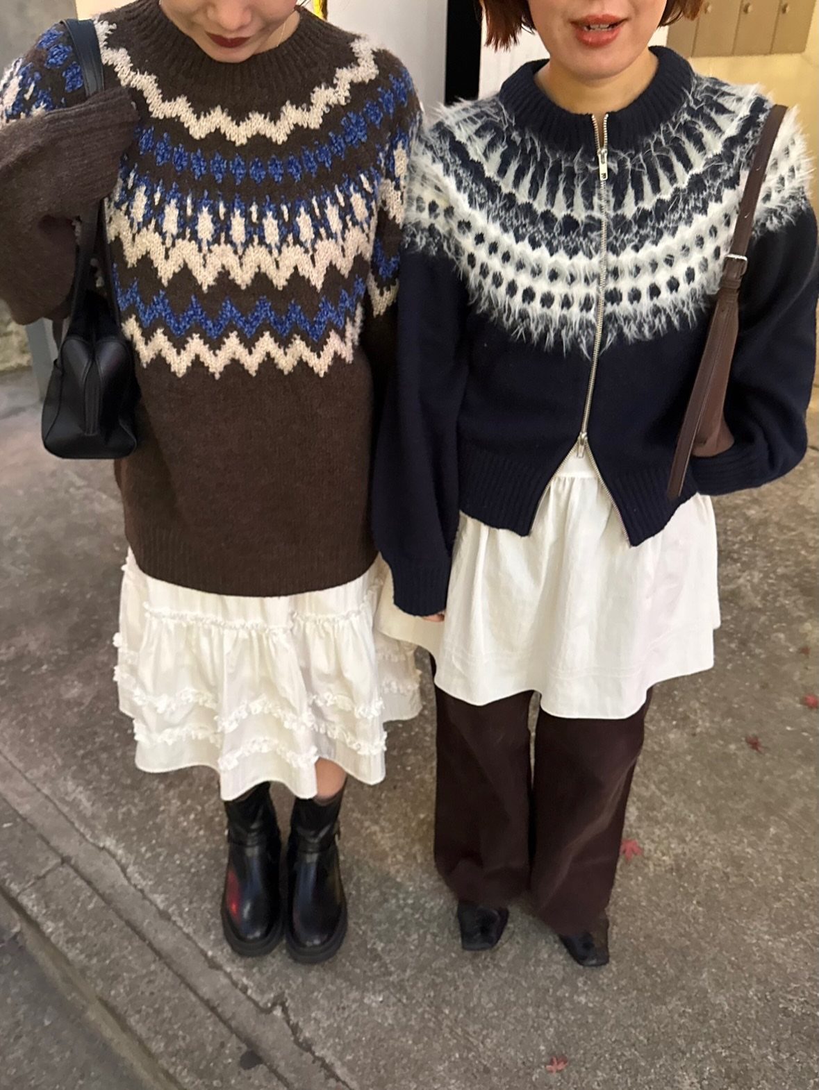 Two women standing on a sidewalk wearing patterned knit sweaters and long skirts.