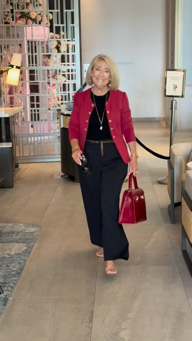 An older woman in a red blazer and black trousers walking through a bright indoor hallway.