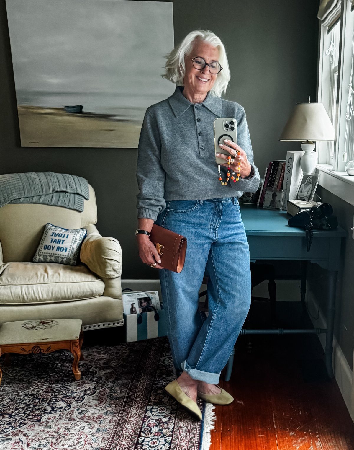 A smiling woman with white hair wearing a grey polo, blue jeans, and beige flats.