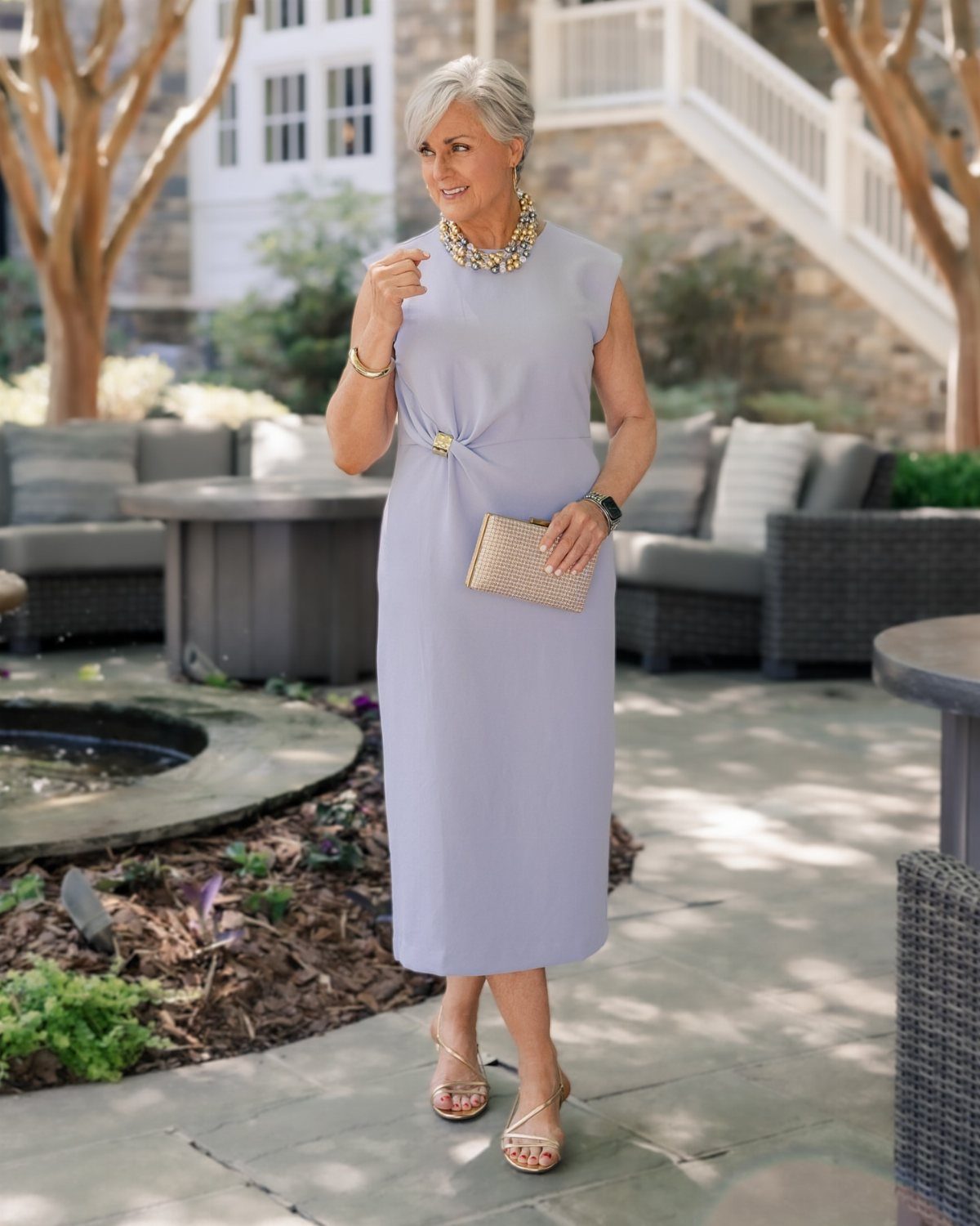 An older woman in a lavender midi dress and gold accessories standing in an outdoor patio setting.