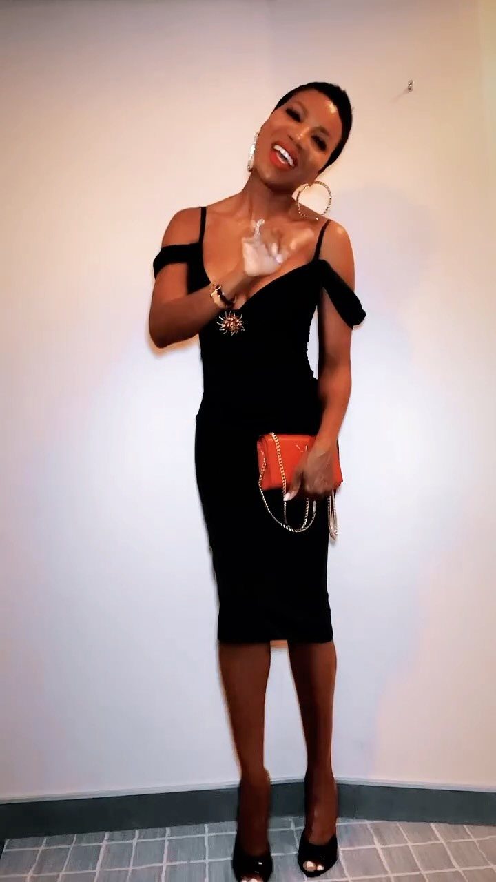 A smiling woman posing in a black off-the-shoulder midi dress and orange clutch.
