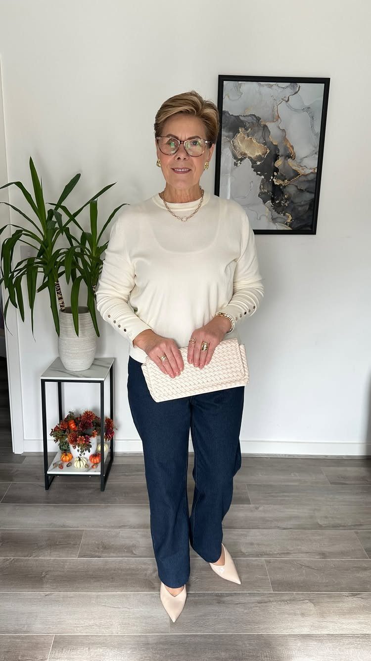 A woman in a cream sweater and navy trousers holding a white clutch bag.