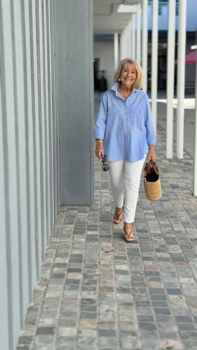 A blonde woman walking outdoors wearing a blue shirt and white trousers.