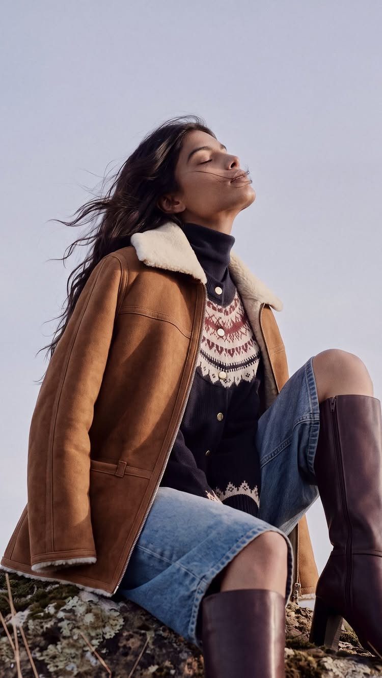 Woman wearing a brown shearling jacket, patterned sweater, denim skirt, and tall brown boots.