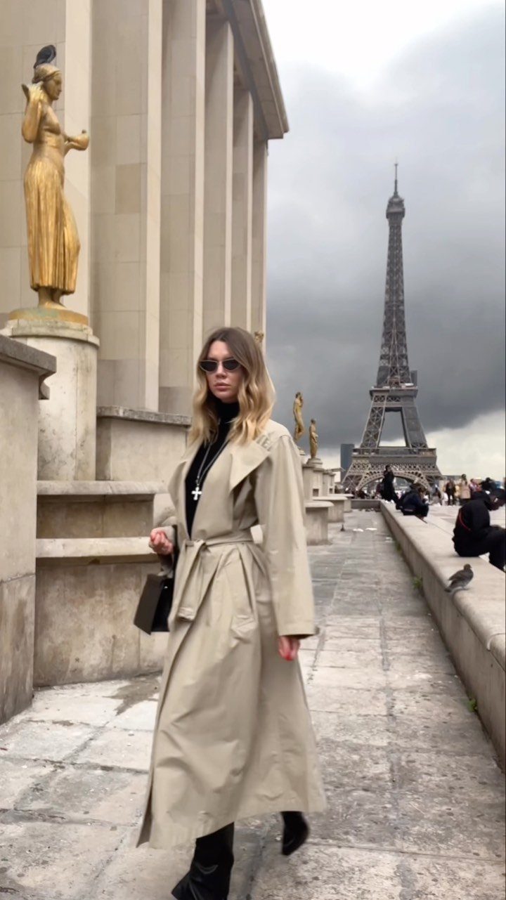 A woman in a beige trench coat walking in Paris with the Eiffel Tower behind her.