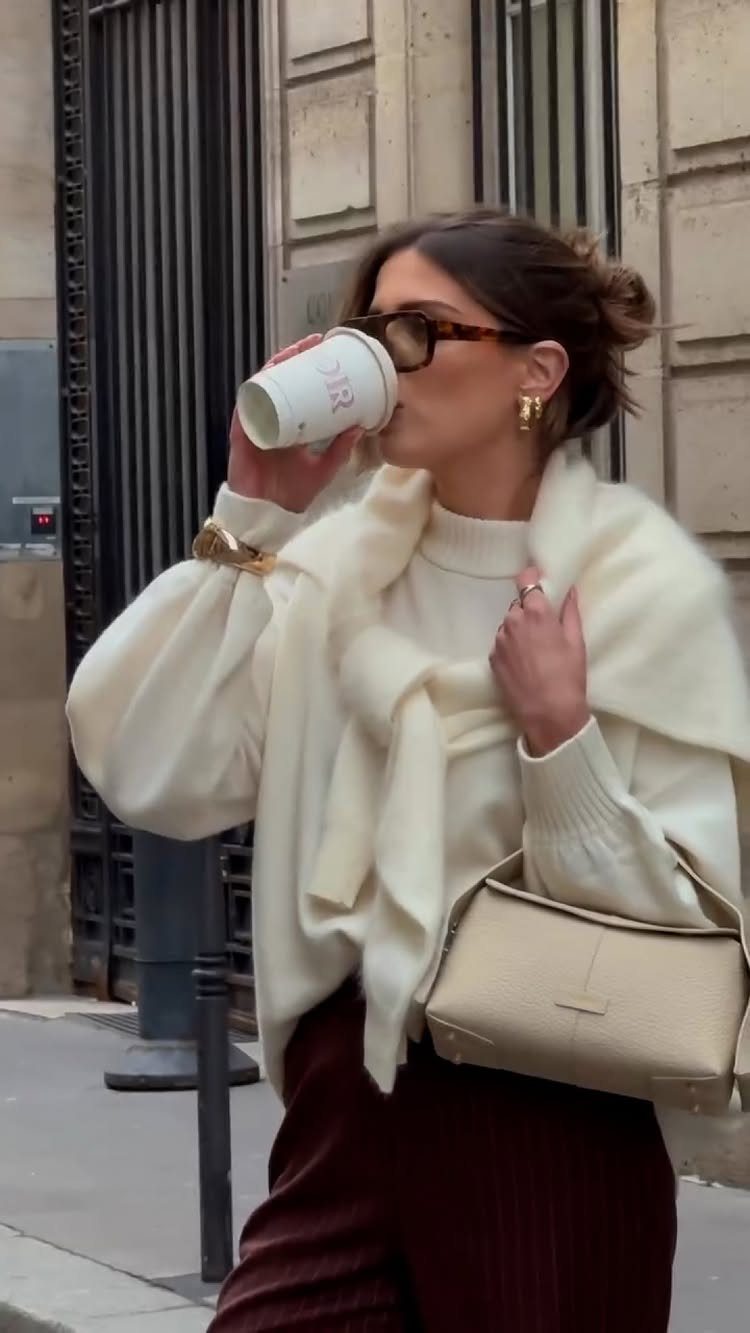 A woman in a white crewneck sweater and cream wrap walking through a city street.