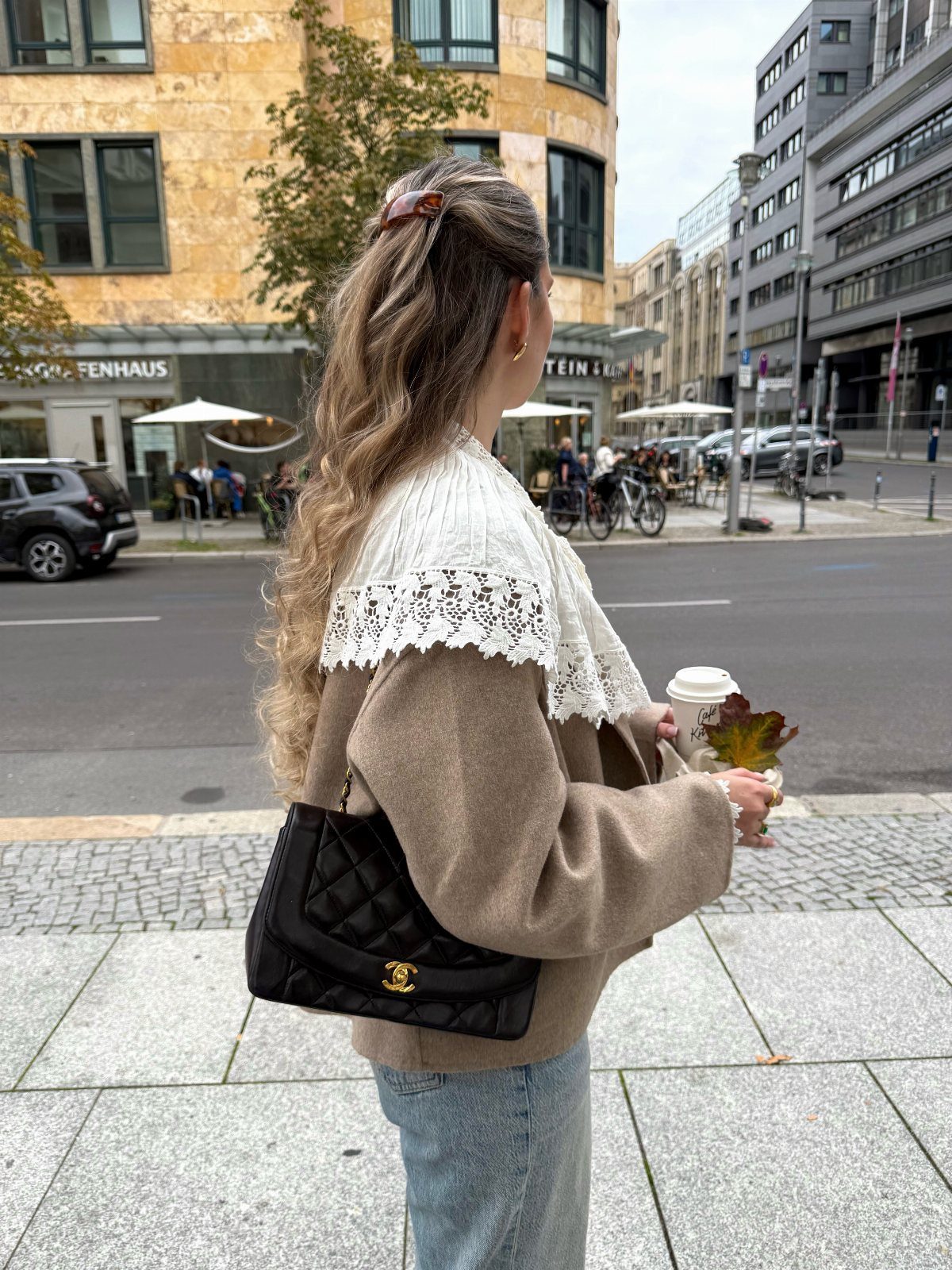 A woman wearing a tan lace-collar sweater and blue jeans walks on a city street.