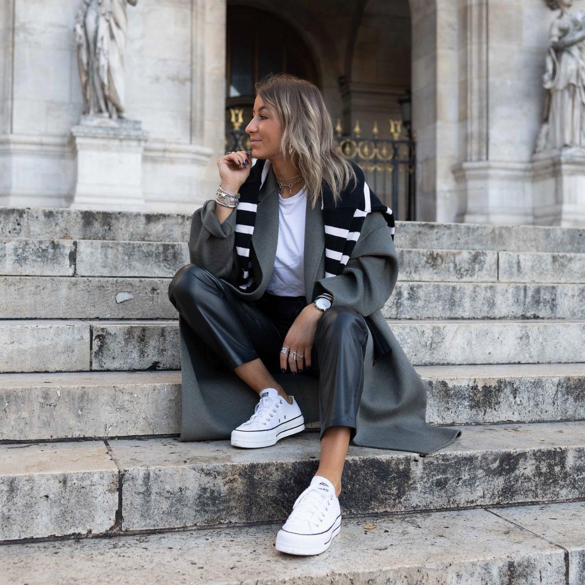 Woman sitting on stone steps wearing a layered fall outfit with leather trousers and white sneakers.