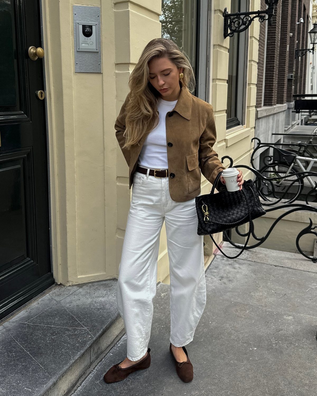 A woman wearing a brown suede jacket, white trousers, and brown loafers standing outdoors.