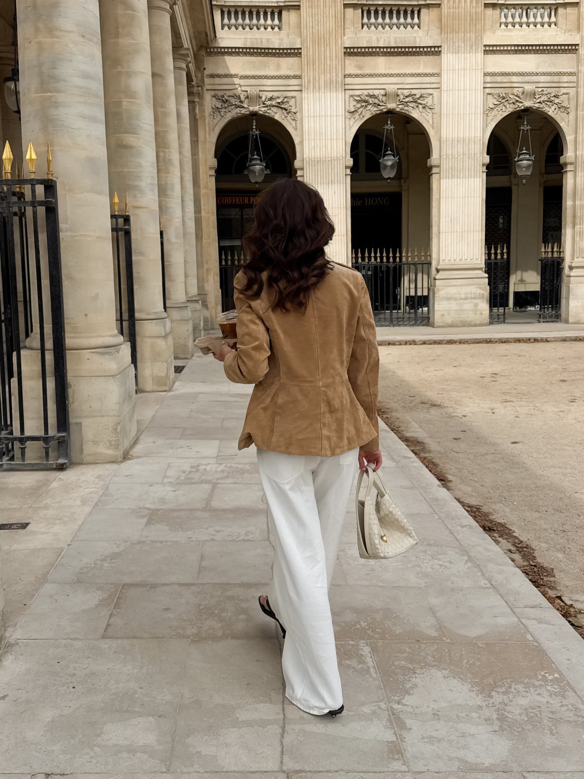Woman in a tan blazer and white trousers walking past classical Parisian architecture.