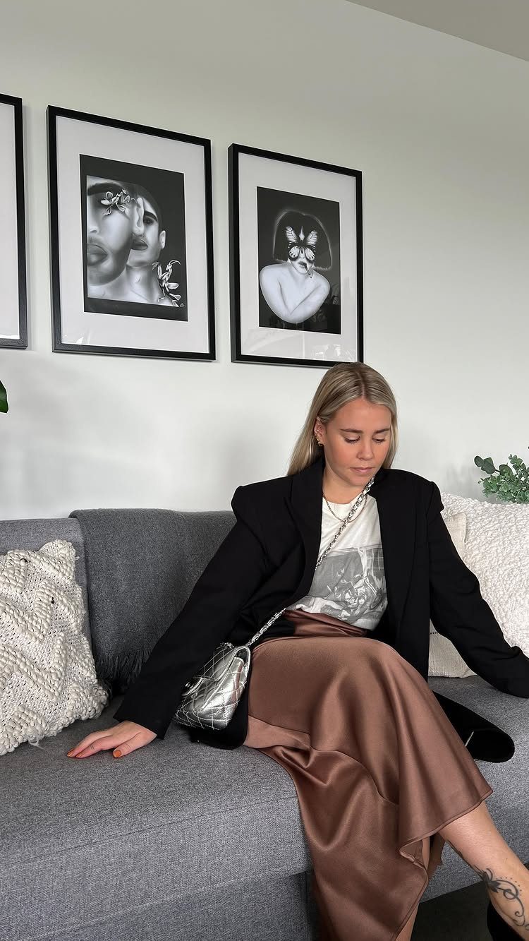 A woman wearing a black blazer, white t-shirt, and brown satin skirt sitting on a sofa.