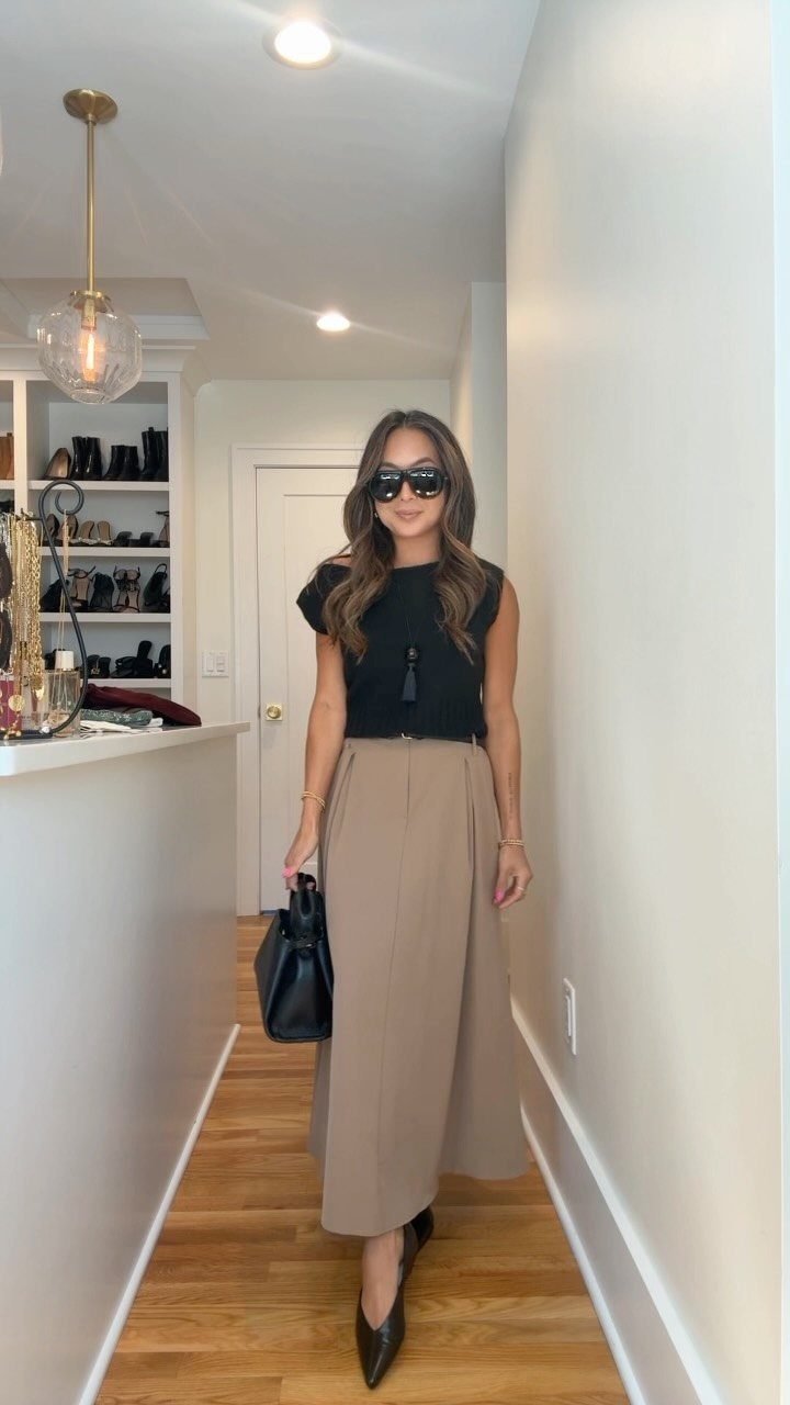 A woman in a black top and tan skirt posing in a hallway wearing sunglasses.