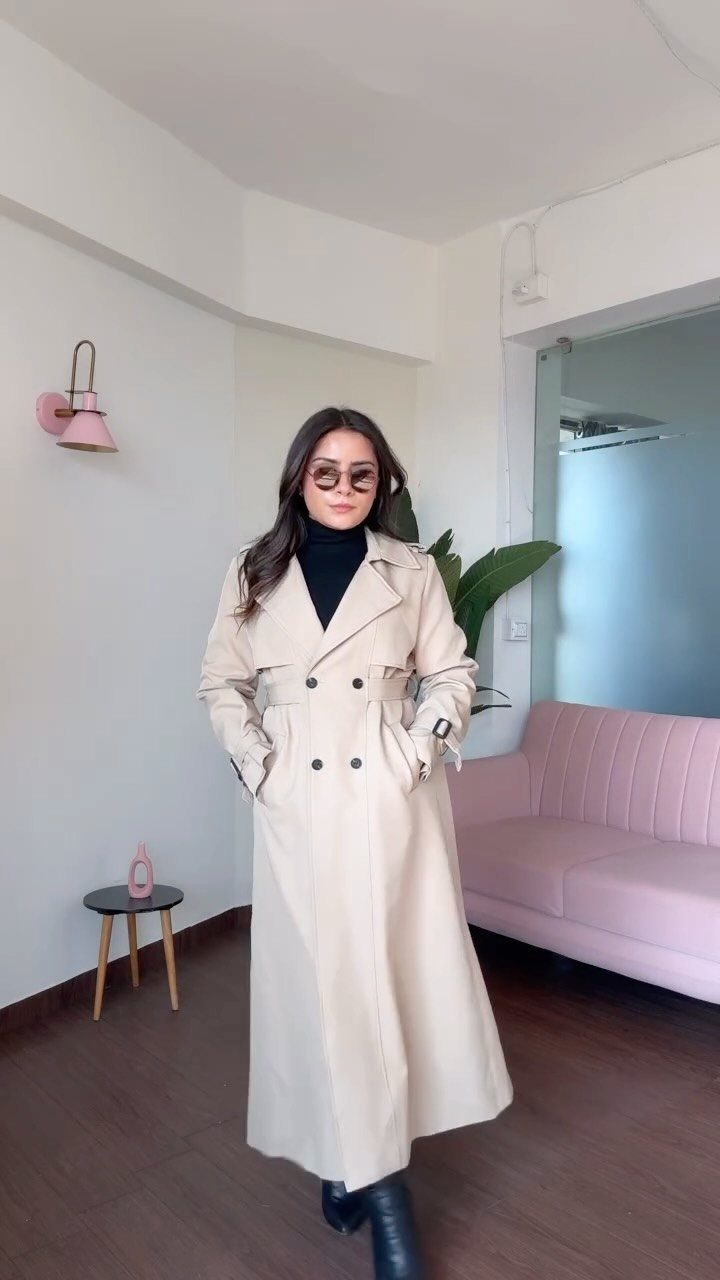 A woman wearing a long beige trench coat and black turtleneck standing in a room.