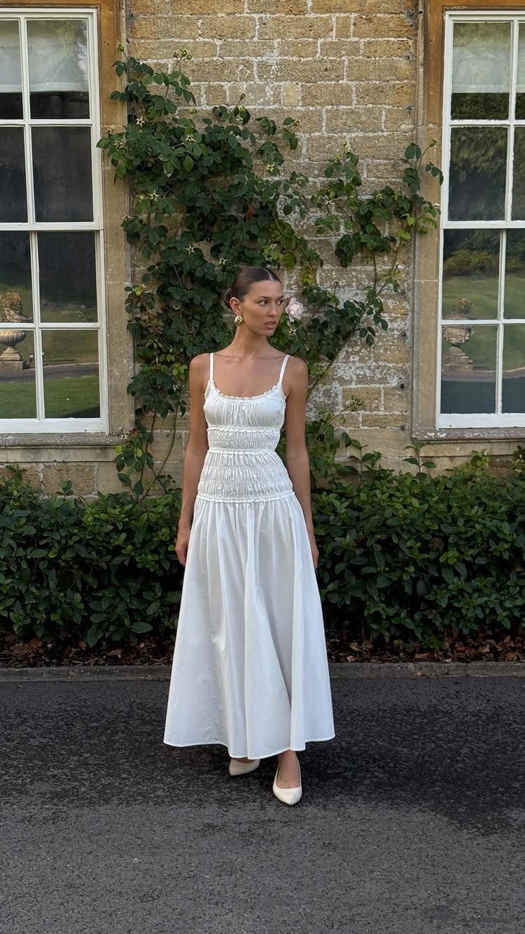 A woman wearing a long white sleeveless breezy dress standing in front of a stone wall.