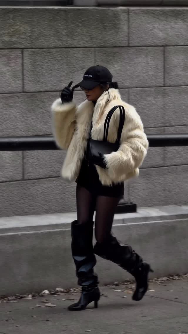 A woman walking in a cream faux fur coat, black cap, and tall black boots.