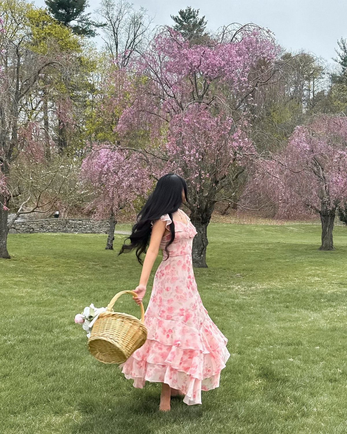 Woman in a pink floral maxi dress walking through a garden with blooming trees.