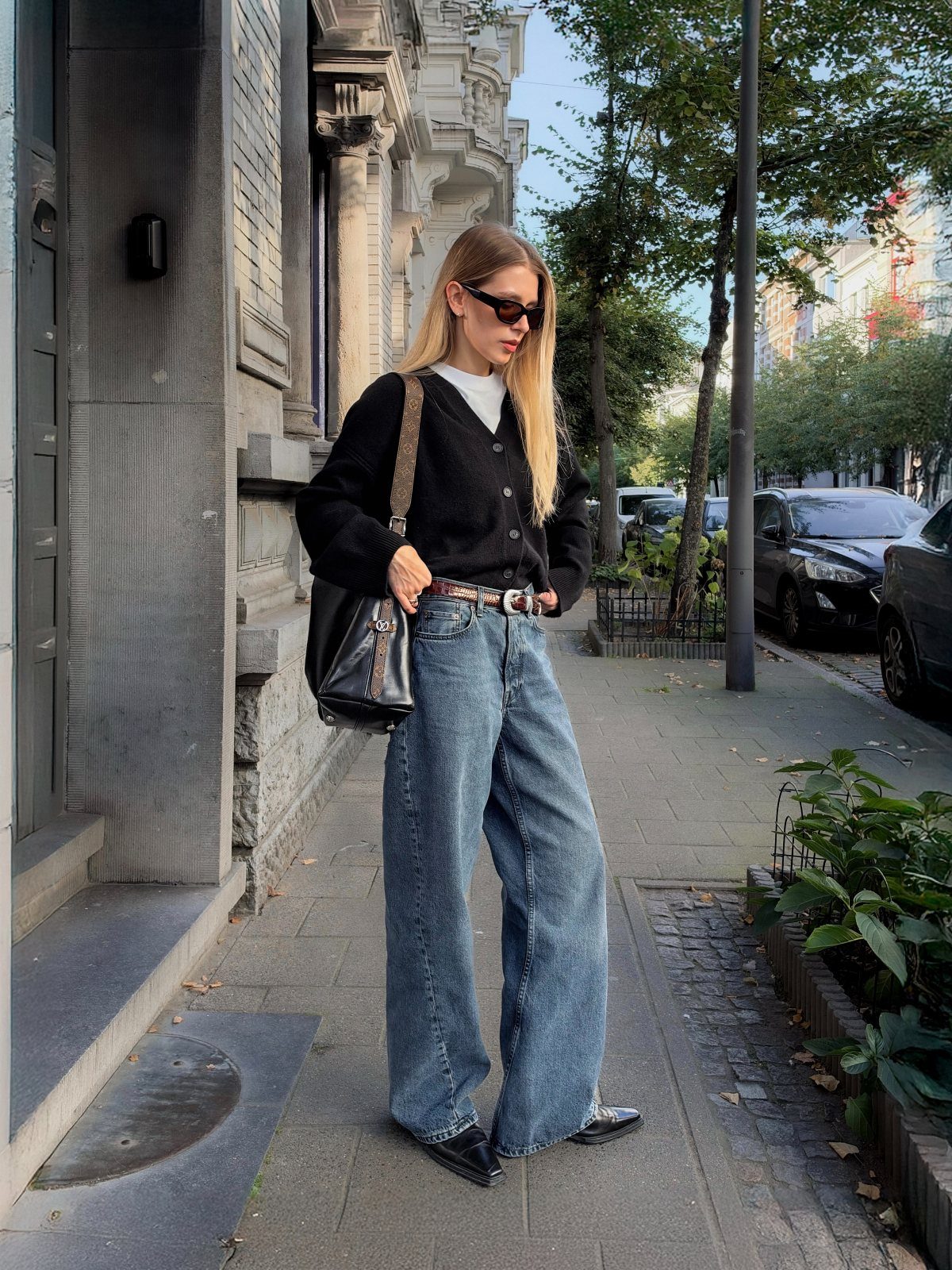 A woman wearing high-waisted wide-leg blue jeans and a black cardigan standing on a city sidewalk.