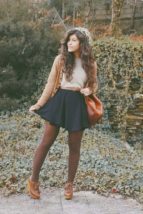 A woman wearing a beige knit sweater, tan cardigan, black skirt, and brown boots outdoors.