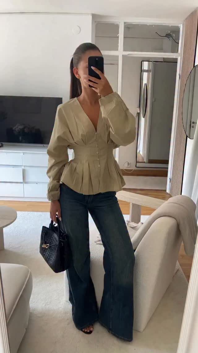 A woman posing in a mirror wearing a beige peplum top and wide-leg denim jeans.