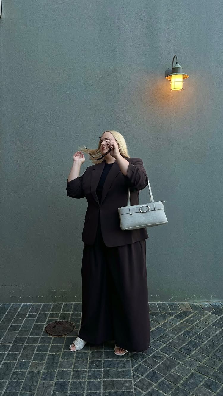 A blonde woman wearing a dark brown blazer, black top, and long skirt holding a white handbag.
