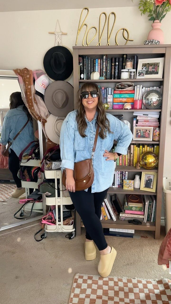 A woman posing in a denim shirt, black leggings, tan clogs, and a brown crossbody bag.