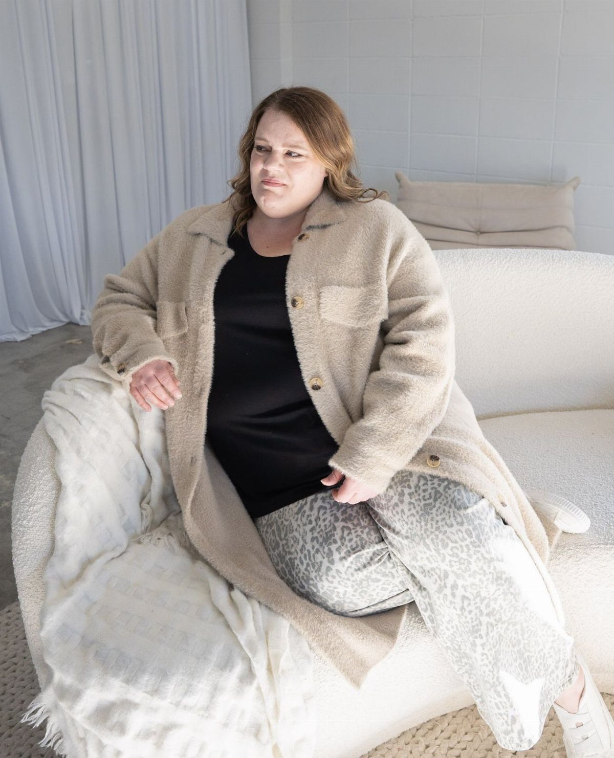 A woman sitting on a white sofa wearing a long beige fuzzy cardigan and patterned pants.