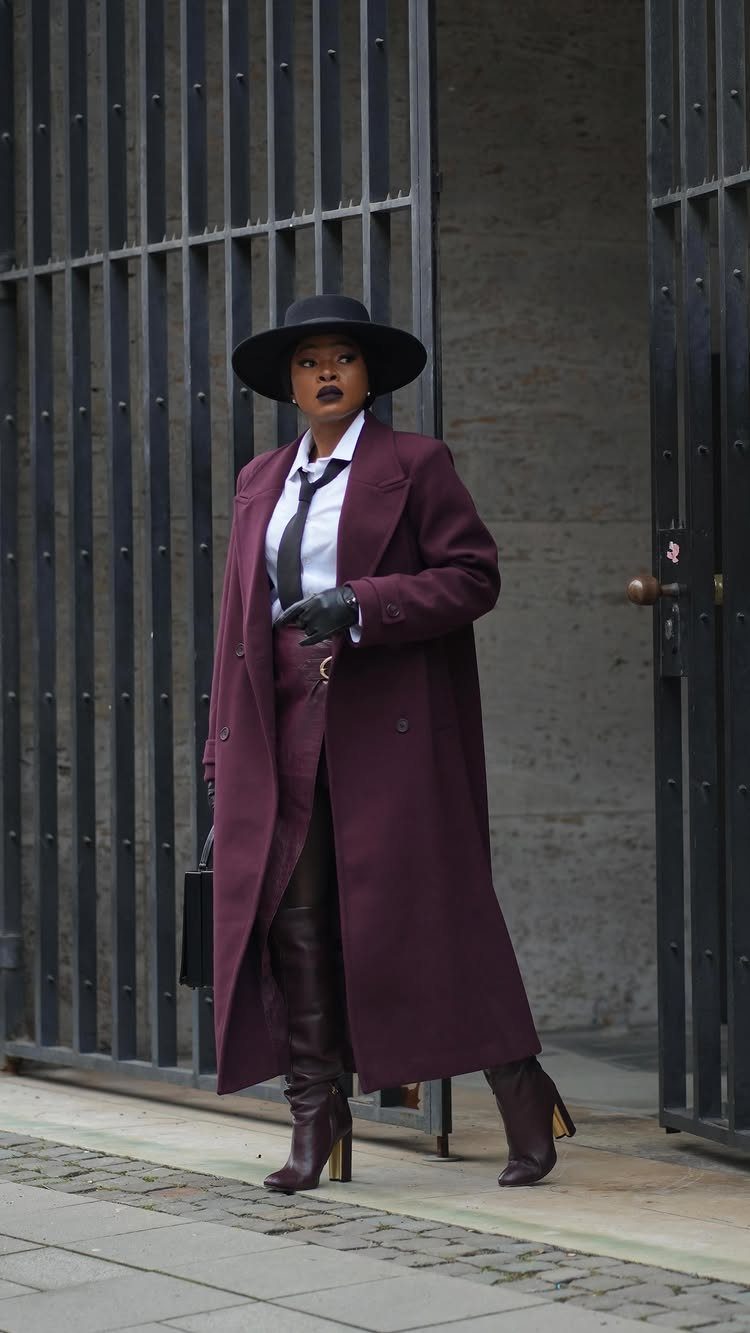 Woman in a long burgundy coat, white shirt, and black tie posing by a metal gate.