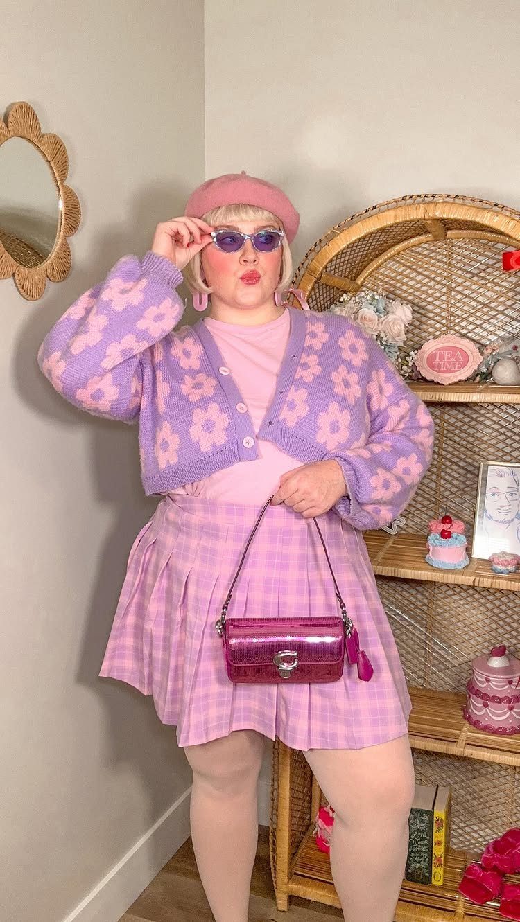 Woman in lavender cardigan and pink plaid skirt holding a small pink handbag.