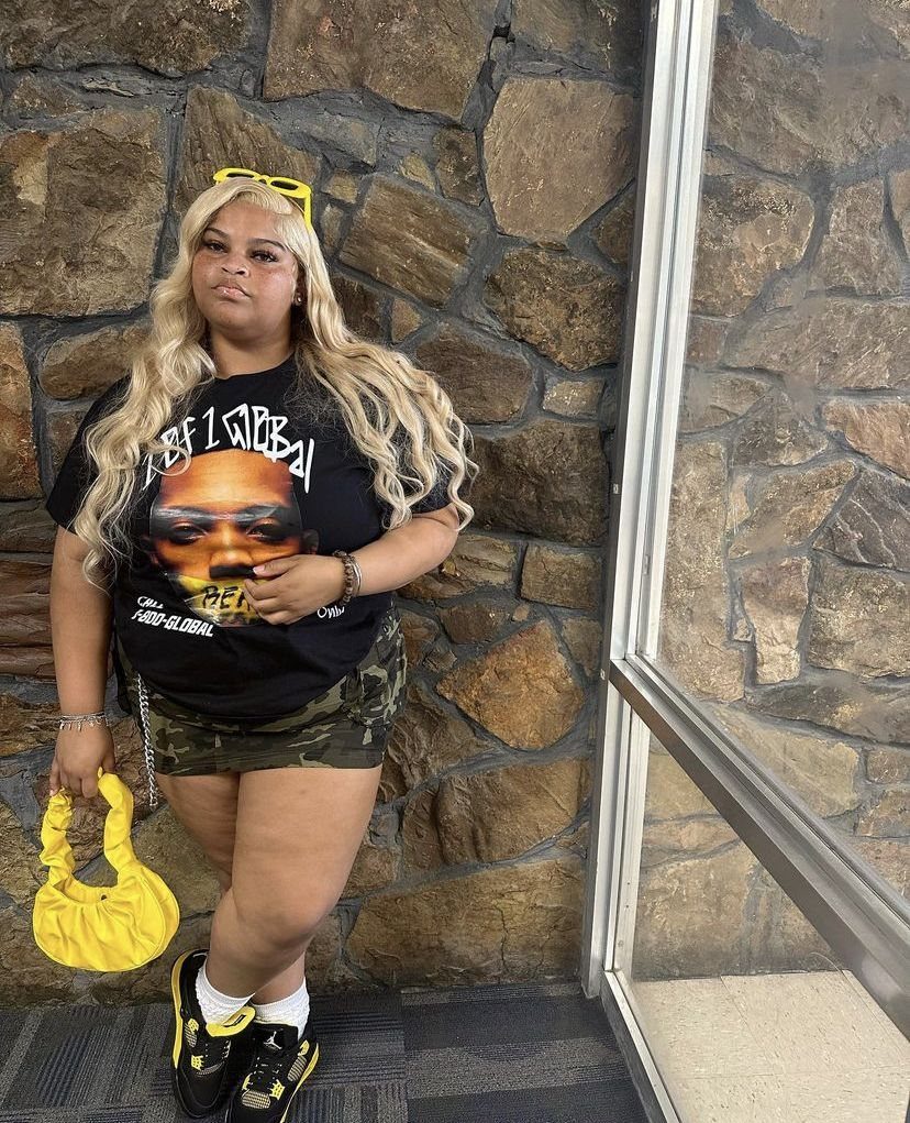 Woman in a black graphic t-shirt and camouflage shorts posing against a stone wall.