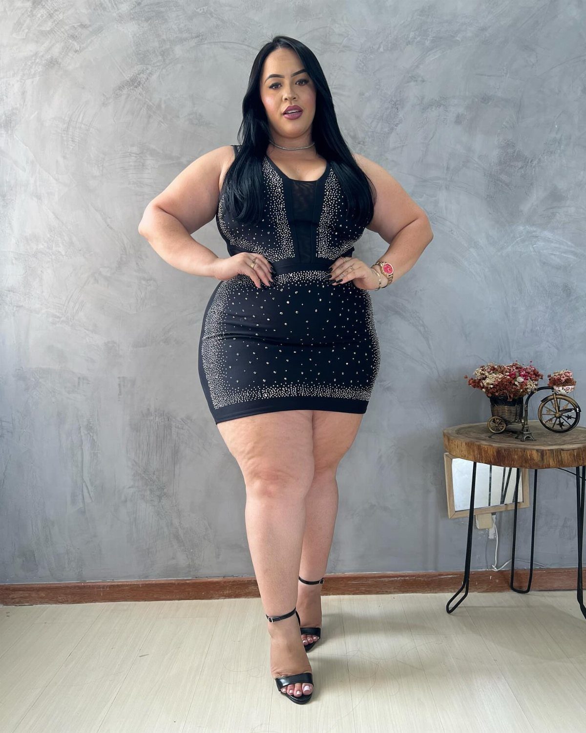 A plus-size woman posing in a black sequined mini dress against a grey wall.