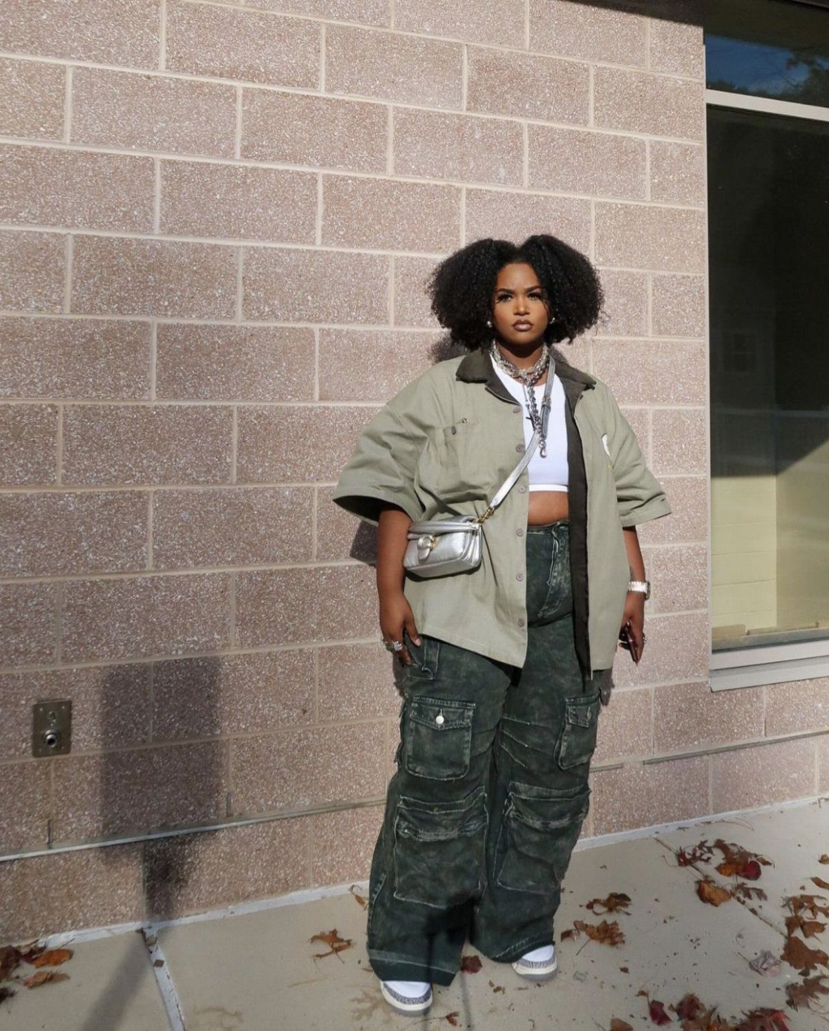 Woman in oversized olive jacket and camouflage cargo pants posing against a brick wall.